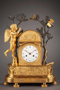 Pendule en bronze ciselé, doré et patiné. Cupidon en équilibre, essaie d'attraper dans son filet un papillon, symbole de l'amour fragile. Son carquois et arc sont posés contre le tronc.