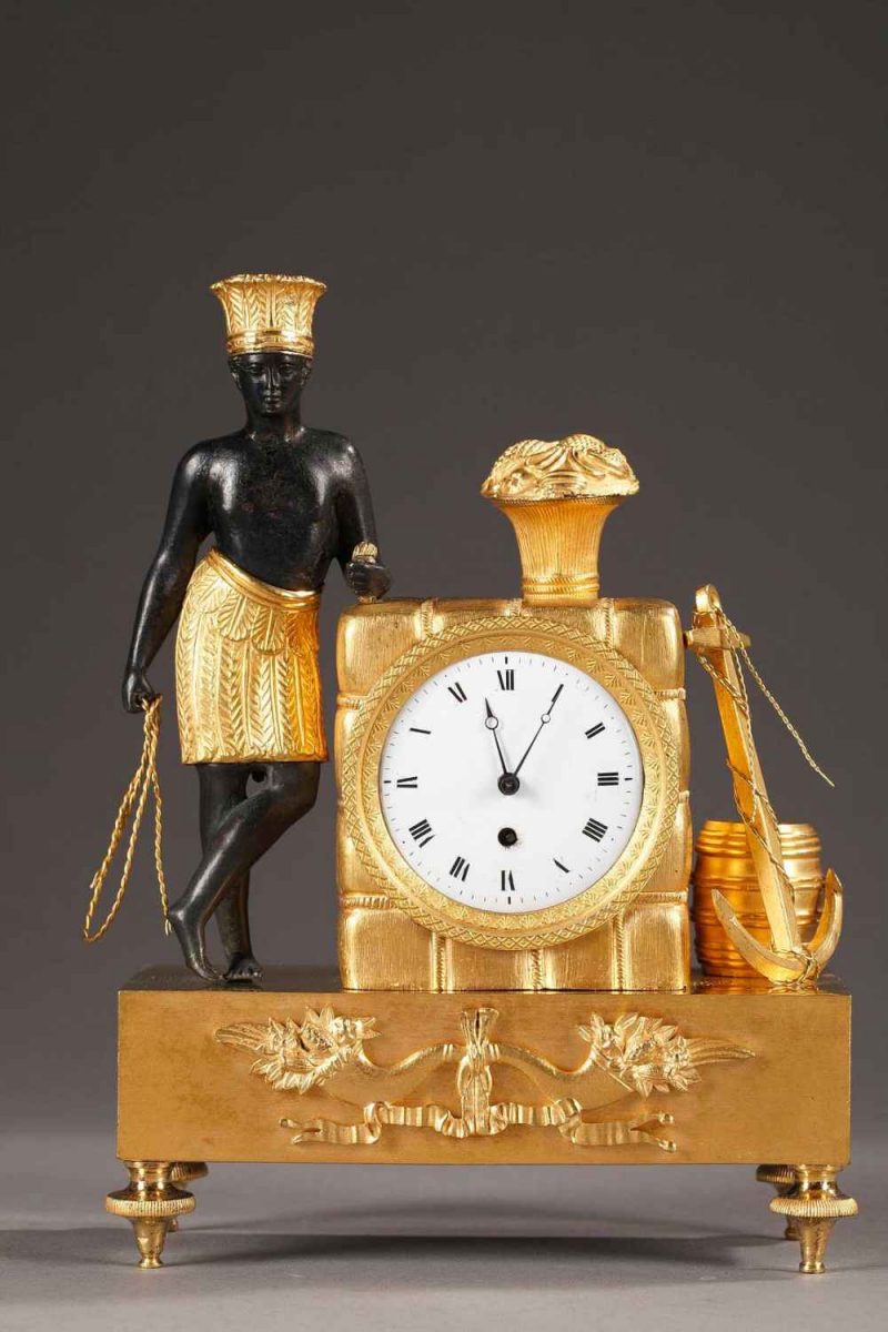 Pendulette en bronze doré, ciselé et patiné. Un africain vêtu d'un pagne et d'une coiffe à plume, est accoudé au ballot dans lequel s'inscrit le mouvement de montre à coq.